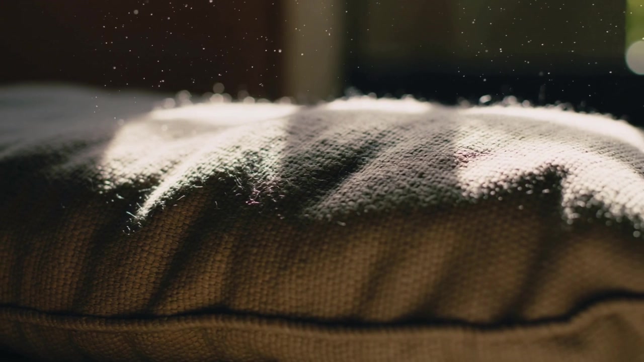 Pulviscolo fluttuante che si alza sopra un cuscino a maglia vicino al telaio di una finestra, luce solare che riscalda la cucitura, bokeh verde.