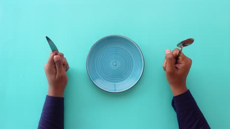 hands holding a fork, knife and spoon next to an empty plate on a blue background