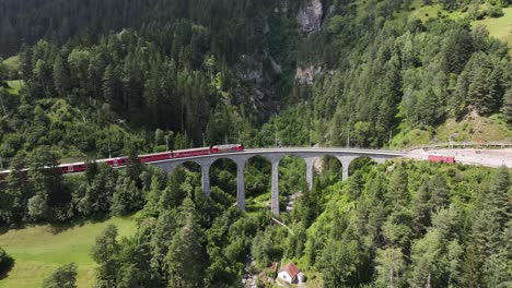 red train crossing a historic stone viaduct in a mountain forest landscape