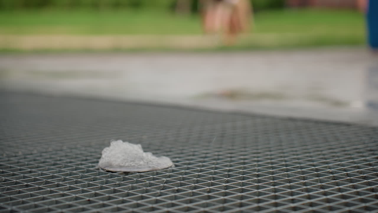 Toddlers standing bare footed near water jet rising from metal grate, summer park setting, gentle splash on grid surface, curiosity and cooling play evident, blurred greenery beyond