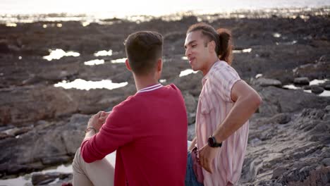 Happy-diverse-gay-male-couple-sitting-on-rocks-and-embracing-at-beach,-slow-motion