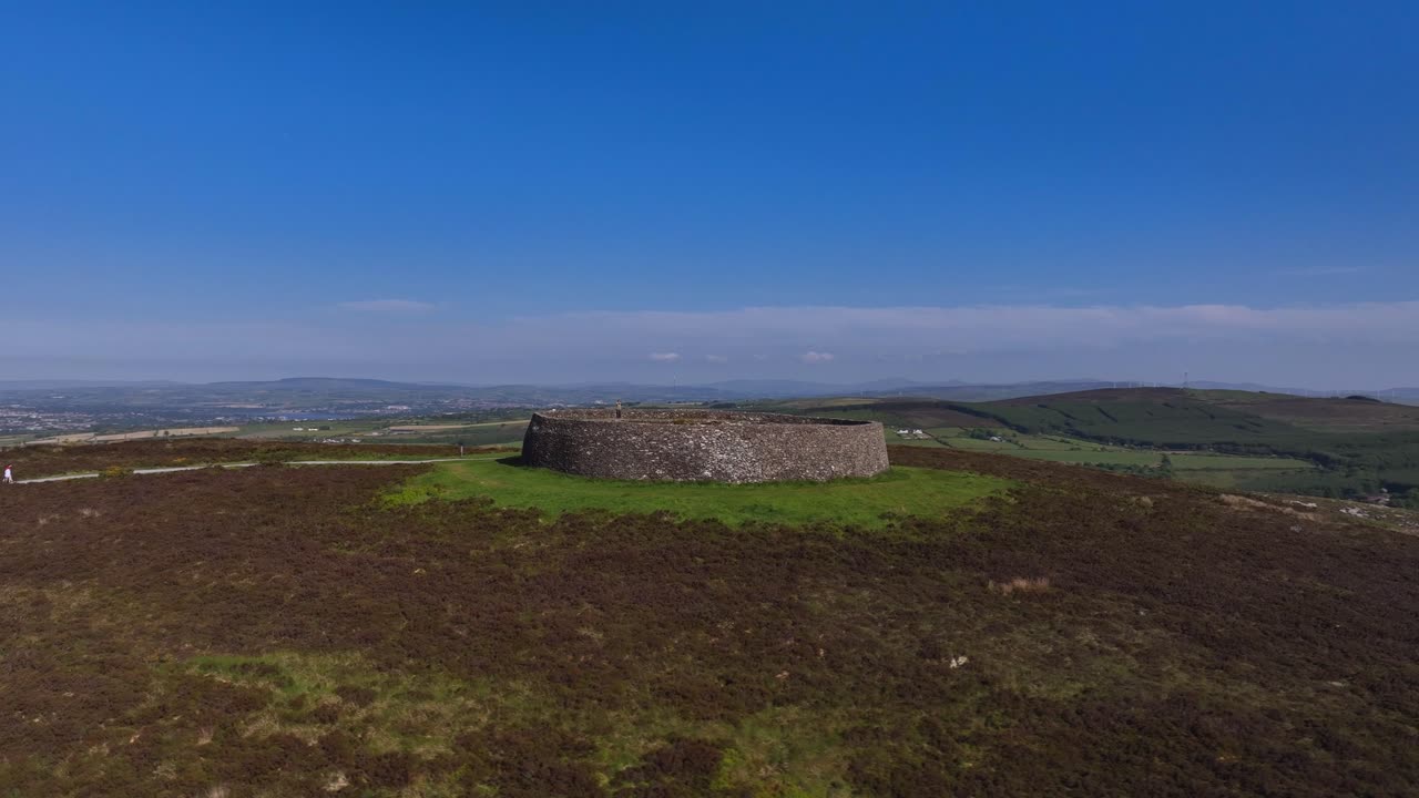 Grianan of Aileach, County Donegal, Ireland, June 2023. Drone orbits clockwise pushing towards the iconic Gaelic Ringfort surrounded by grass and wild brown shrubs with Lough Swilly in the distance.