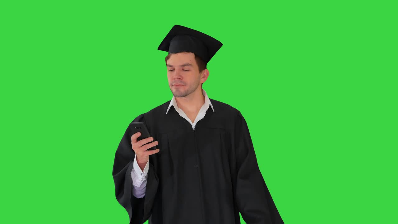 Male student in graduation gown checking his phone while walking on a Green Screen, Chroma Key