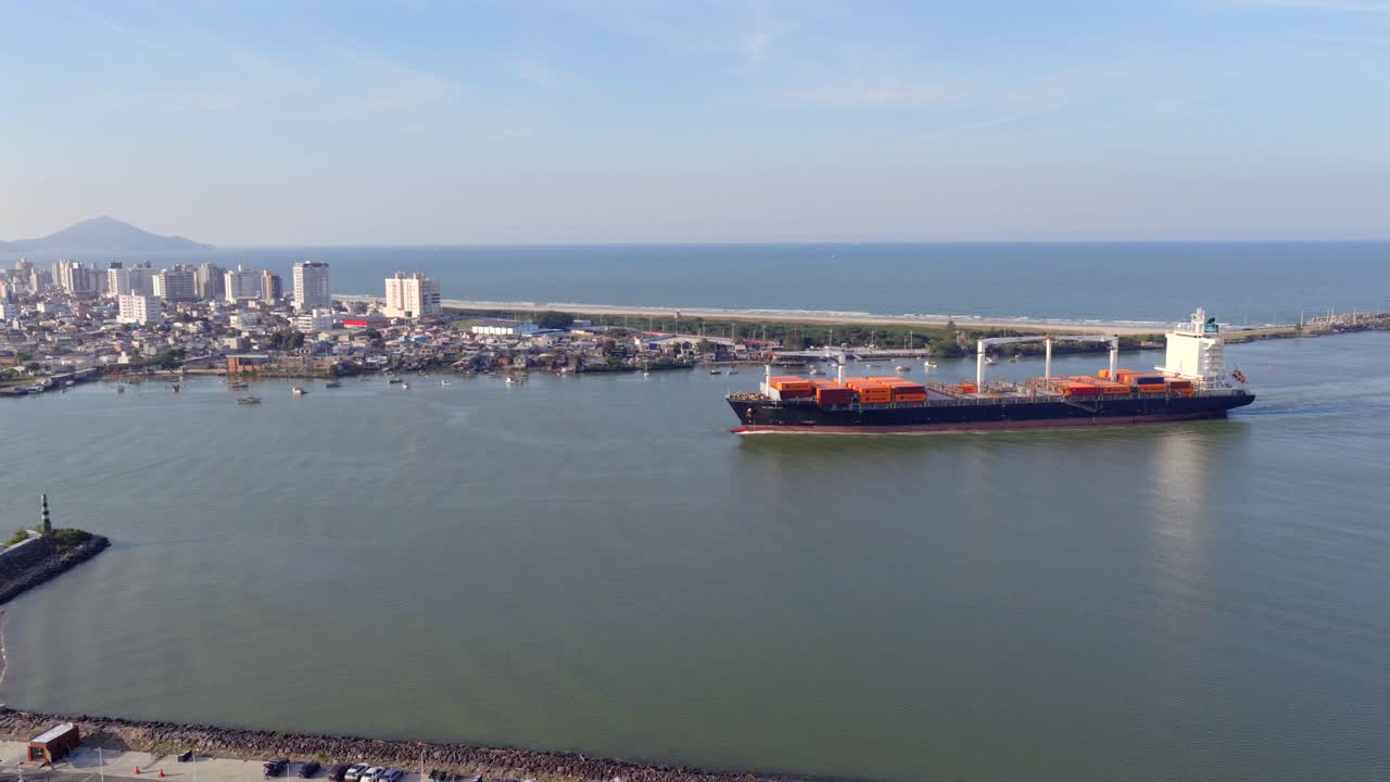 Drone captures massive transport vessel entering river mouth from open ocean, with coastal buildings near and flat horizon during clear day