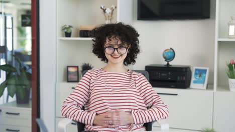 a portrait of a beautiful young curly girl sitting at a table with a laptop in a spacious light office by handing hair and glasses for vision and smiles at the camera