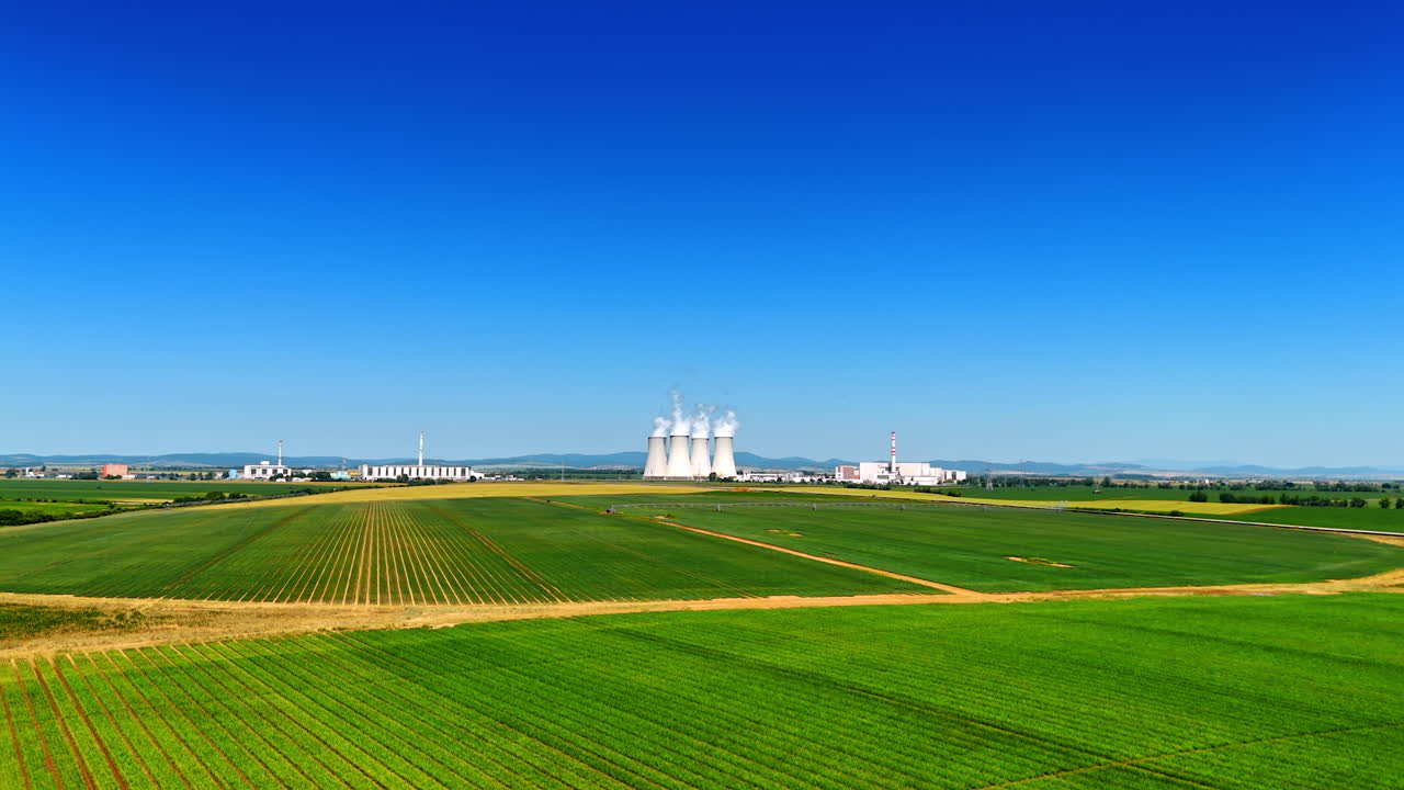Footage over the green fields in the countryside. Wide pipes of the industrial plant produce white smoke. Slovakia industry