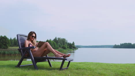 Beautiful-brunette-in-a-swimsuit-lying-on-a-sun-lounger-on-the-lake-smiling-working-remotely-via-the-Internet-in-the-business-sphere.-The-concept-of-country-life