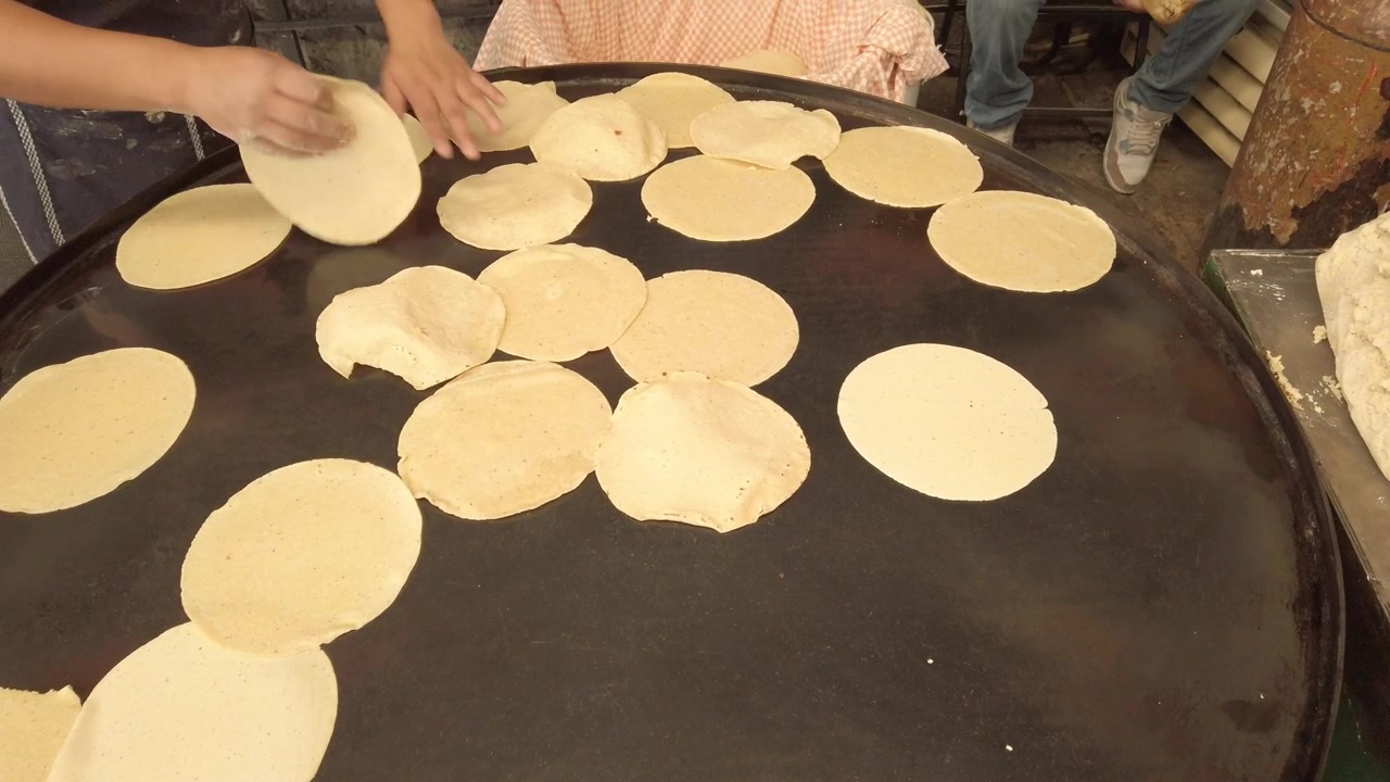 Close view of workers preparing fresh tortillas by hand on large heated griddle inside busy traditional Mexican food cooking area