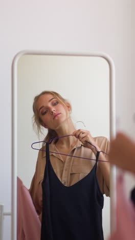 young woman with ponytail looks at reflection in mirror choosing between elegant pink and classic black dress for evening party slow motion