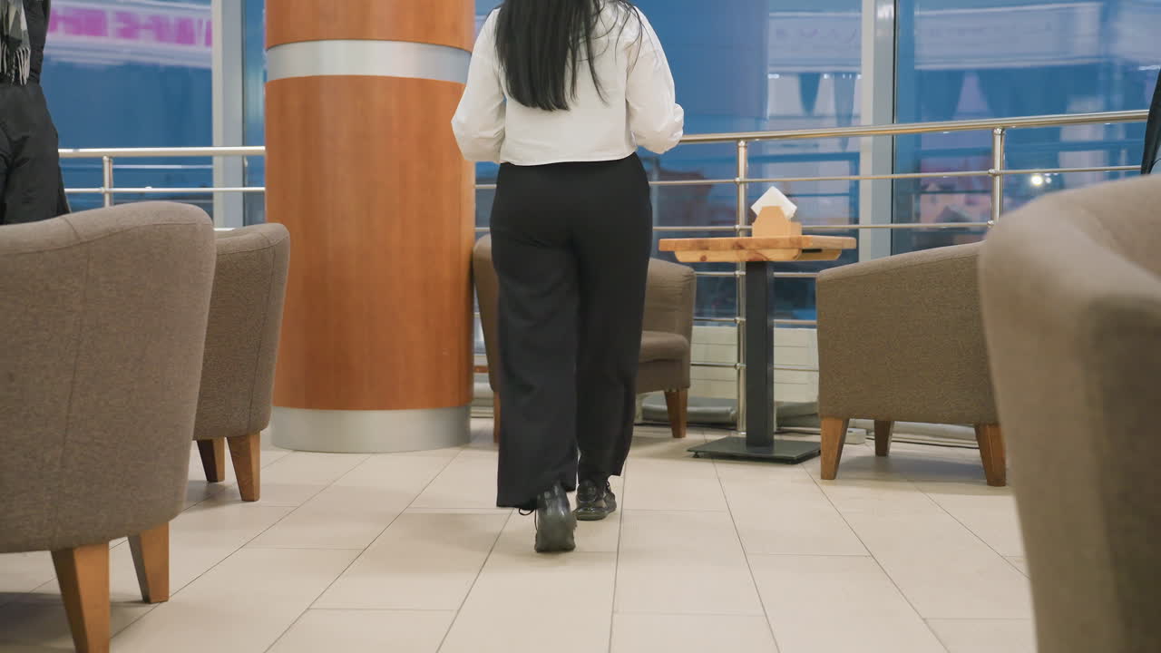 Young woman walks up to empty seat inside cozy eatery, gently places coffee cup and gadget on wooden table before sitting down with legs crossed and relaxing while looking around interior