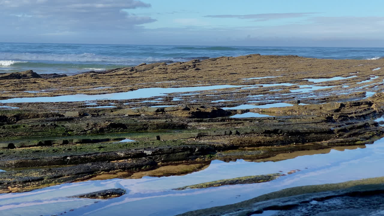 The rocky patterns of the Sagres coast shimmer under sunlight, shaped by centuries of ocean tides Jagged rocks rise against rolling Atlantic waves under clear Sagres skies, a scene of wild energy