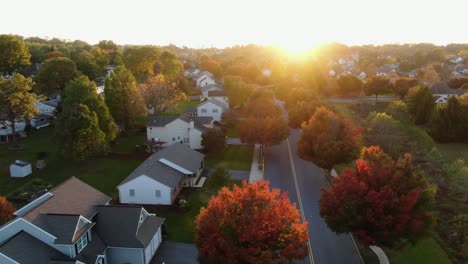 sunset starburst highlights autumn neighborhood scene with homes and colorful trees, cars and trucks on street at intersection, aerial drone shot