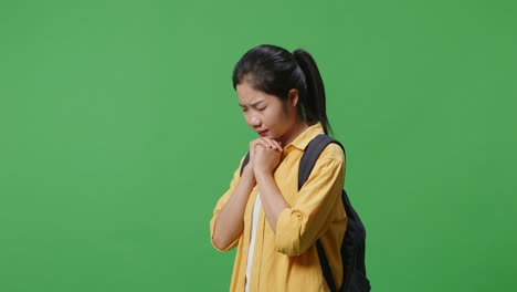 side view of asian woman student with a backpack closing her eyes prays for something while standing in the green screen background studio