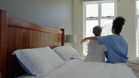 rear view of female doctor and senior woman with arm around sitting on bed in a comfortable home 4k