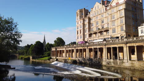 Static-Shot-of-Pulteney-Weir-and-the-Empire-Hotel-in-Bath,-Somerset-on-Beautiful-Summer’s-Morning-with-Blue-Sky-with-Birds-Flying-Through-Frame