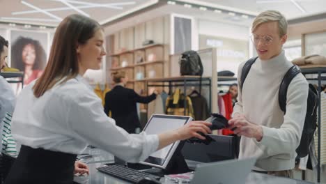 clothing store: young man at checkout counter buys clothes paying with contactless nfc smartphone, friendly retail sales assistan packs merchandise. contemporary fashion shop with of designer brand