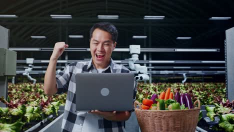 feliz agricultor masculino asiático con canasta de verduras celebrando usando una computadora portátil en una granja inteligente