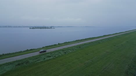 drone shot of a lone car driving along a long straight dike next to a wide body of water on a cloudy day.