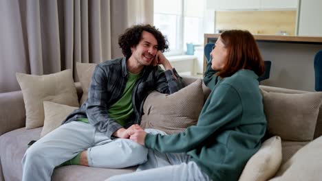 Happy-guy-with-curly-hair-laughs-loudly-and-has-fun-while-communicating-with-his-brunette-girlfriend-while-sitting-on-the-sofa-at-home-in-a-modern-apartment