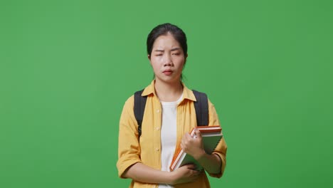 불만족한 아시아 여성 학생은 배과 몇 권의 책을 가지고 카메라를 바라보고 녹색 스크린 배경 스튜디오에 서있는 동안 머리를 흔들고 있습니다.