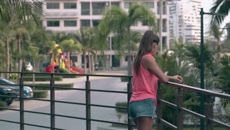 trendy-girl-in-bright-top-stands-by-fence-and-looks-at-palms