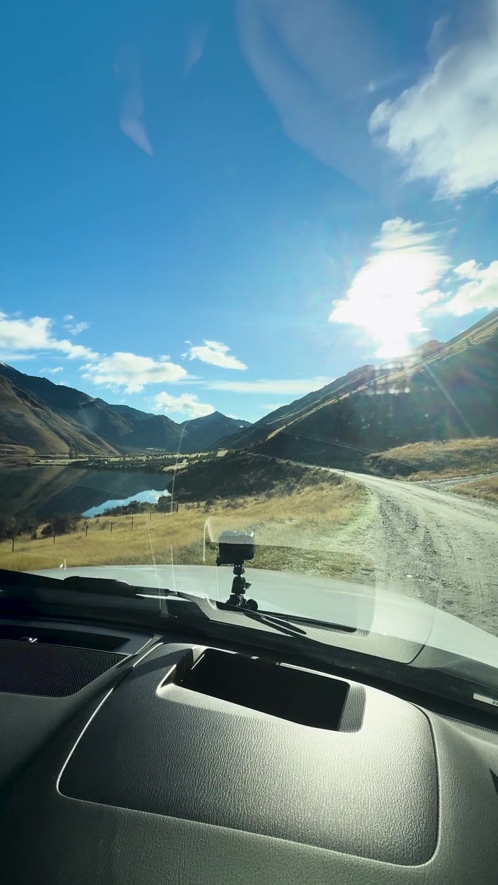 Il tergicristallo dell'auto si muove sul parabrezza, rivelando un paesaggio montano illuminato dal sole e un lago in pieno giorno.