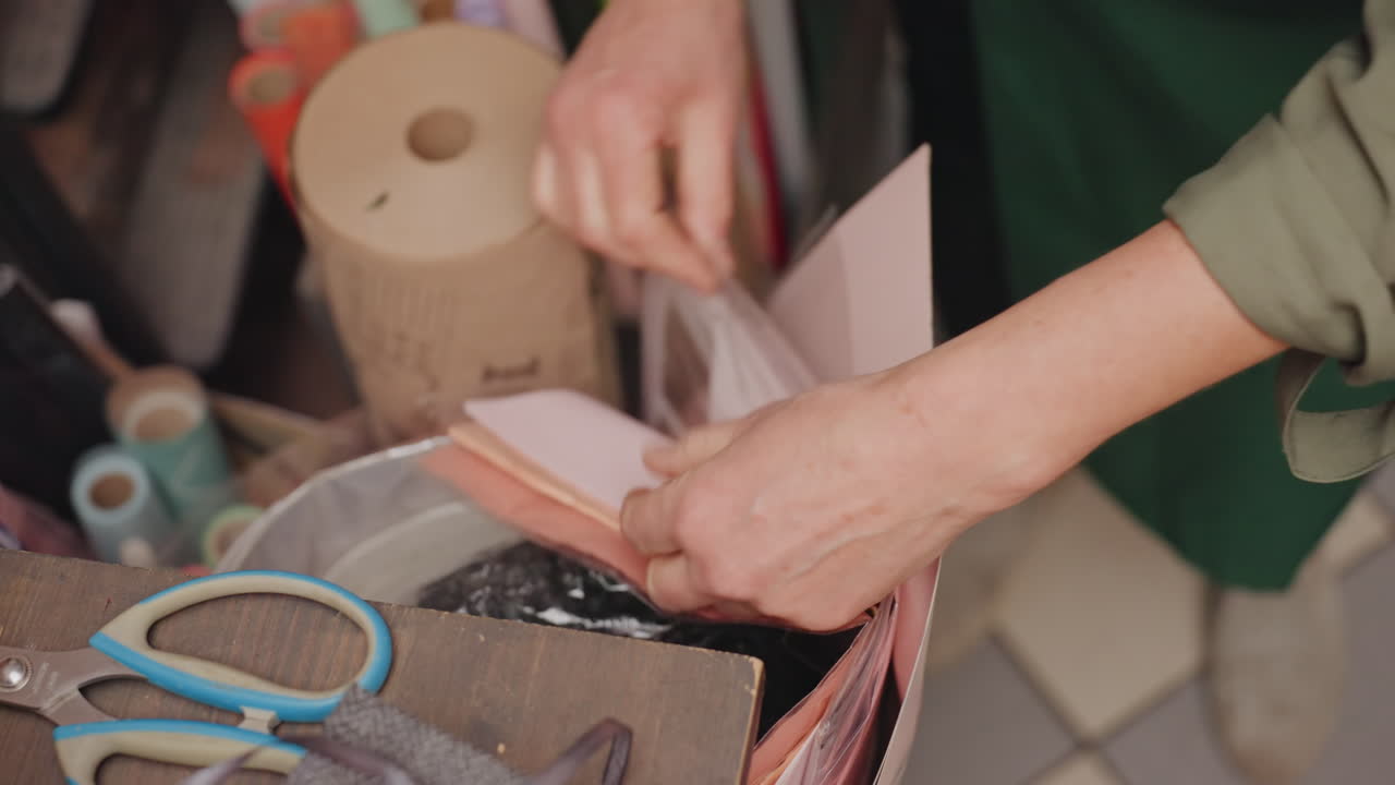 Mani di fiorista caucasica che avvolgono carta rosa attorno ai gambi; forbici e nastro adesivo su bancone piastrellato. Piegatura e cordonatura dettagliate, sequenza attenta delle fasi di imballaggio per bouquet da consegna, destrezza costante.