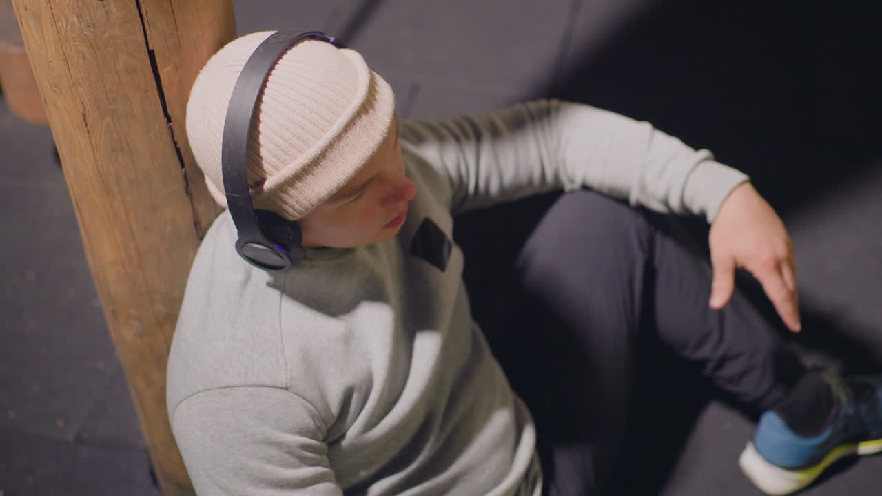 Ung mand sidder ved en søjle og lytter til musik iført hue og sweater, lænet op ad en betonsøjle i et solbeskinnet hjørne af byen, afslappet kropsholdning og afslappede sneakers, indadvendt stemning med blød