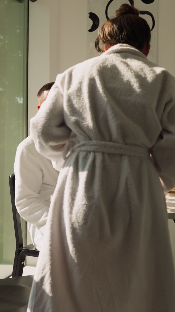 una mujer se une a un hombre sentado en la mesa de la cocina. la novia en bata de baño de terry viene al novio sentado a la mesa para compartir el té de la mañana juntos. rutinaria de la mañana