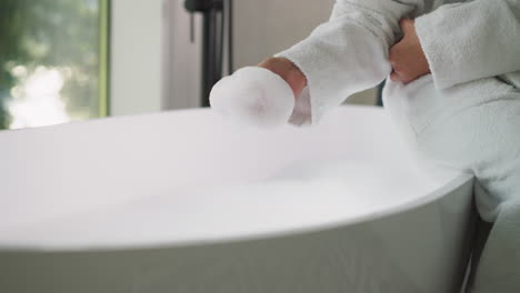 Woman takes foam sitting on bathtub edge. Young lady in soft bathrobe plays with bubble bath preparing for body care procedures at home. Home relaxation
