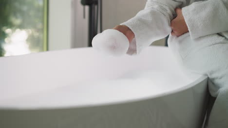 woman takes foam sitting on bathtub edge. young lady in soft bathrobe plays with bubble bath preparing for body care procedures at home. home relaxation