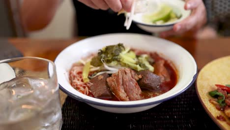 A person garnishes a bowl of rice vermicelli in Northern Thai curry sauce with pickled vegetables at a restaurant table in Bangkok. Natural daylight, close-up, steady camera