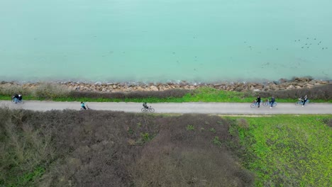 People-riding-bicycles-near-ocean-shore-in-Loix-village-in-Île-de-Ré-Island-France,-Aerial-orbit-around-shot