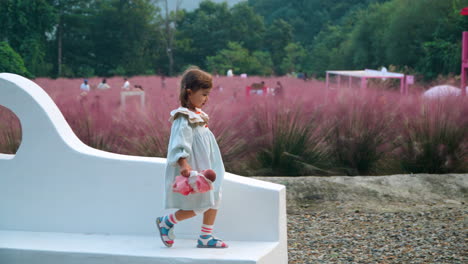 Pink-Muhly-Herb-Island-Park-–-Verspieltes-Kleinkind-Läuft-Auf-Einer-Steinigen-Bank,-Hält-Eine-Spielzeugpuppe-In-Der-Hand-Und-Springt-In-Zeitlupe-Herunter-–-Ökotourismus-In-Südkorea
