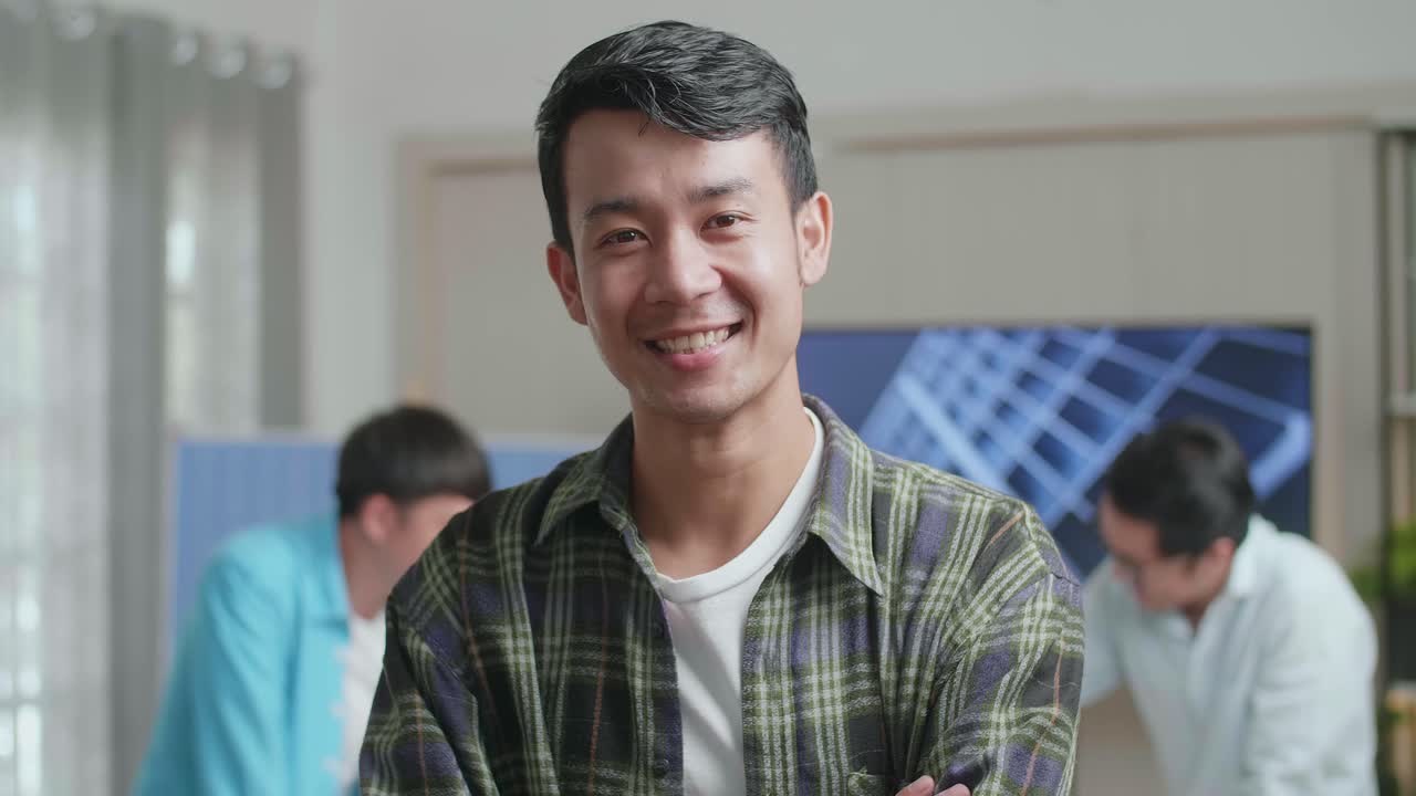 미소 짓는 아시아 남성 엔지니어가 동료들이 사무실에서 태양전지 작업에 대해 논의하는 동안 카메라에 팔을 고 있습니다.