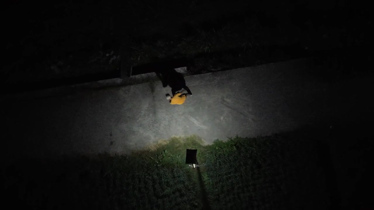 Slow motion drone camera descends and rotates providing a top-down aerial view of a man shadow boxing at night. He wears a cap and trains on a dark path illuminated by a bright spotlight.