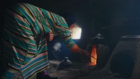 wide shot of a moroccan woman at a fireplace baking traditional bread inside clay made house at a rural village in the atlas mountains.