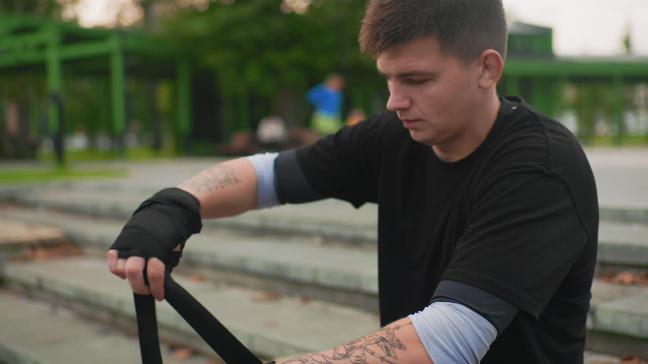 Giovane uomo bianco che avvolge le mani su gradini di cemento, avambracci tatuati, maglietta nera e manicotto al gomito, espressione concentrata, parco urbano sullo sfondo, luce serale, movimenti ravvicinati che mostrano attenzione