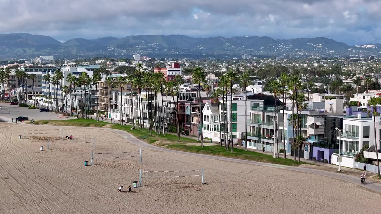 Et filmisk 70 mm luftdroneoptag, der langsomt roterer fremad over Venice Beach, og fanger ejendomme langs Ocean Front Walk i Los Angeles, Californien, med Santa Monica-bjergene i baggrunden.
