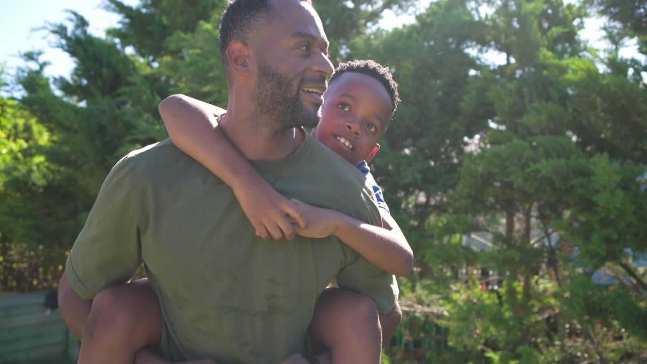 Pai negro dando carona nas costas ao filho num quintal ensolarado, enquanto a criança aponta para a esquerda e o pai se vira sorrindo.