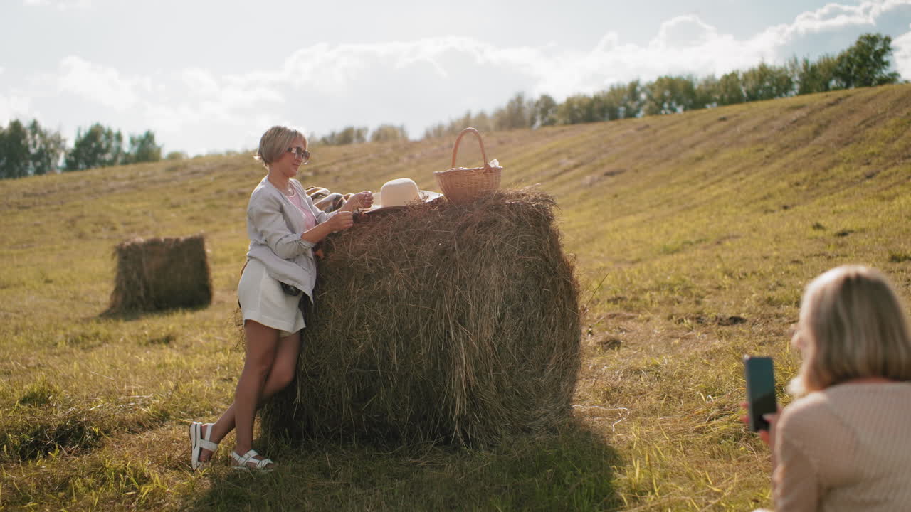 Eine Frau hockt und macht Fotos von einer stilvollen Freundin, die sich die Haare richtet, während sie sich in sonnenbeschienener Landschaft an einen Heuballen lehnt, ein entspannter ländlicher Moment mit Picknickkorb, goldenem Licht und friedlicher offener Landschaft.