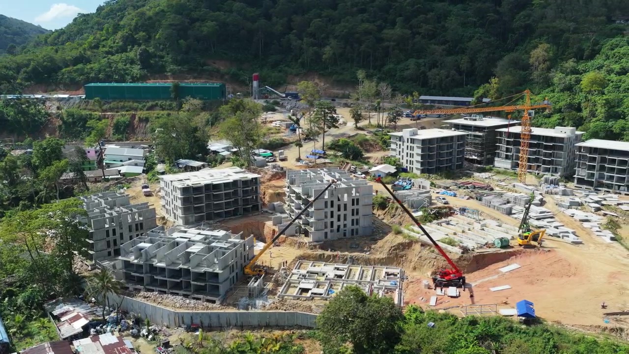 Pengambilan gambar udara pembangunan gedung yang aktif di Phuket dikelilingi perbukitan tropis, tinjauan orbit drone menara derek tinggi