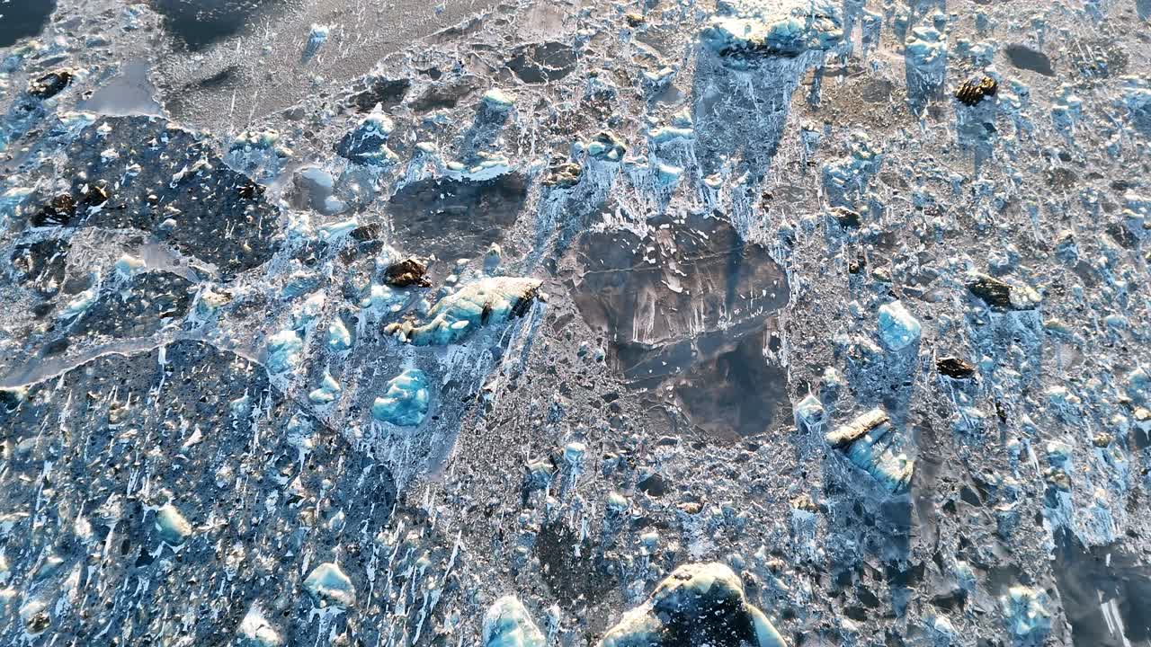 Drone shot of cracked icy surface with scattered blue ice chunks in Iceland. Sunlight reflects on rugged texture, revealing natural patterns and frozen details across glacial landscape.