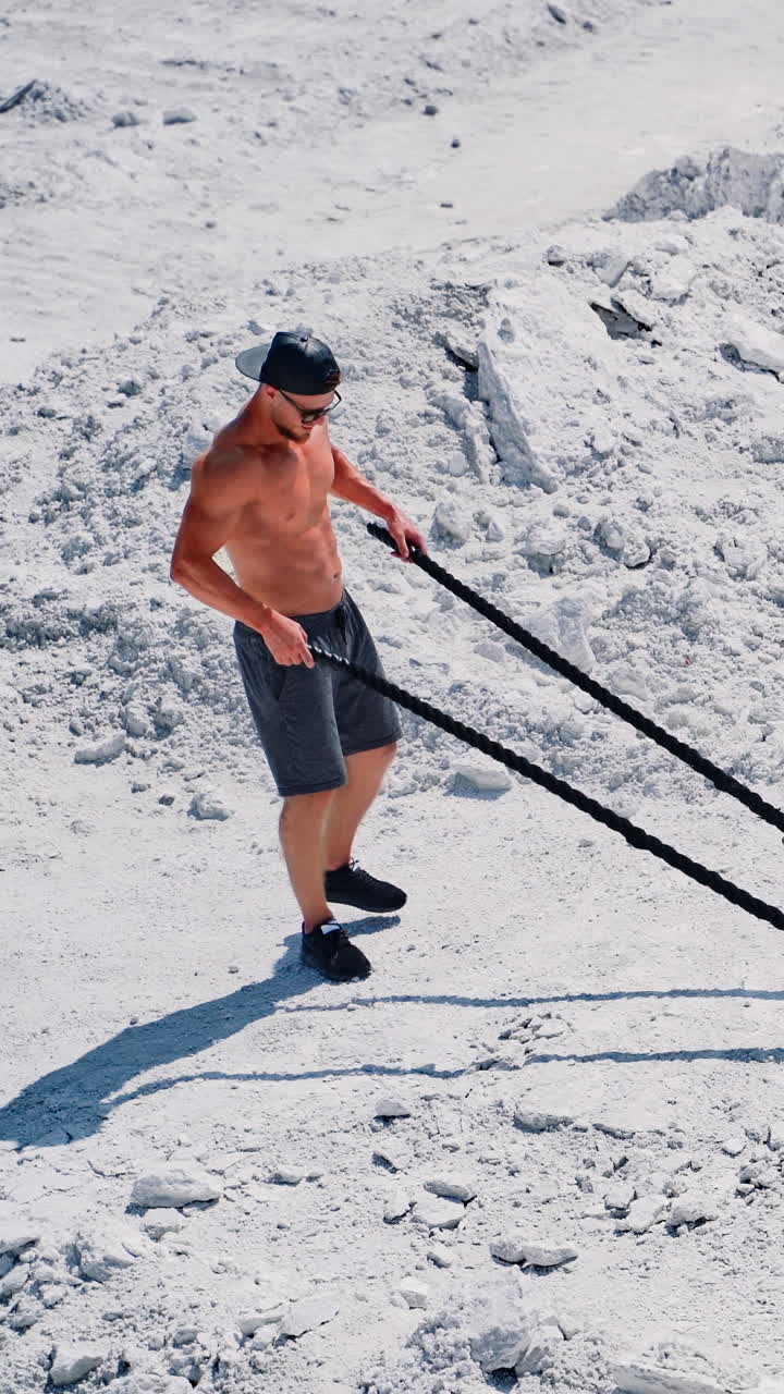fitness athlete in a cap and sunglasses prepares for training with ropes outdoor. Heavy training in a quarry. Intense workout with ropes in white mountains. Strong attractive bodybuilder. Lifestyle. White landscape. Vertical video
