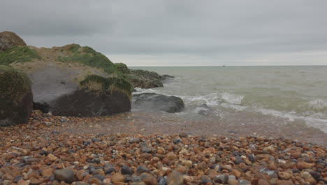 Tiro-Bajo-De-Una-Playa-De-Guijarros-En-Inglaterra-En-Un-Día-De-Nubes,-Ola-Rompiendo-Cerca-De-La-Cámara