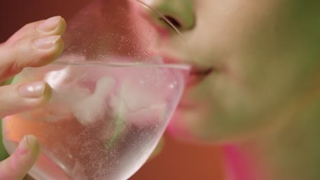 Close-up-shot-of-a-beautiful-woman-with-full-beautiful-lips-drinking-a-fresh-glass-of-water-with-ice-cubes-on-hot-day-to-quench-her-thirst-against-orange-background-with-yellow-purple-contrast-in-face