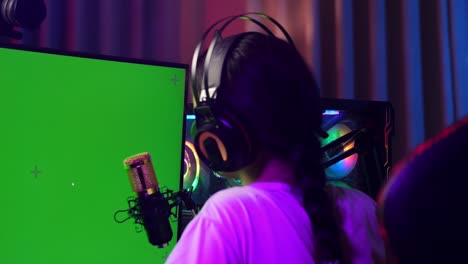 close up back view of asian girl streamer using green screen personal computer, live stream waving hand and talking on the microphone.  desk illuminated by rgb led strip light