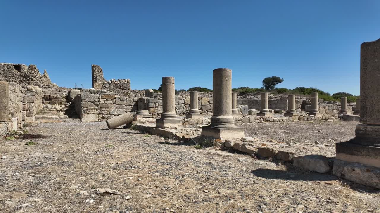 Stabilized handheld shot walking through the Roman ruins of King Juba II's palace in Lixus. The footage features ancient stone columns and walls under a blue sky in Larache, Morocco