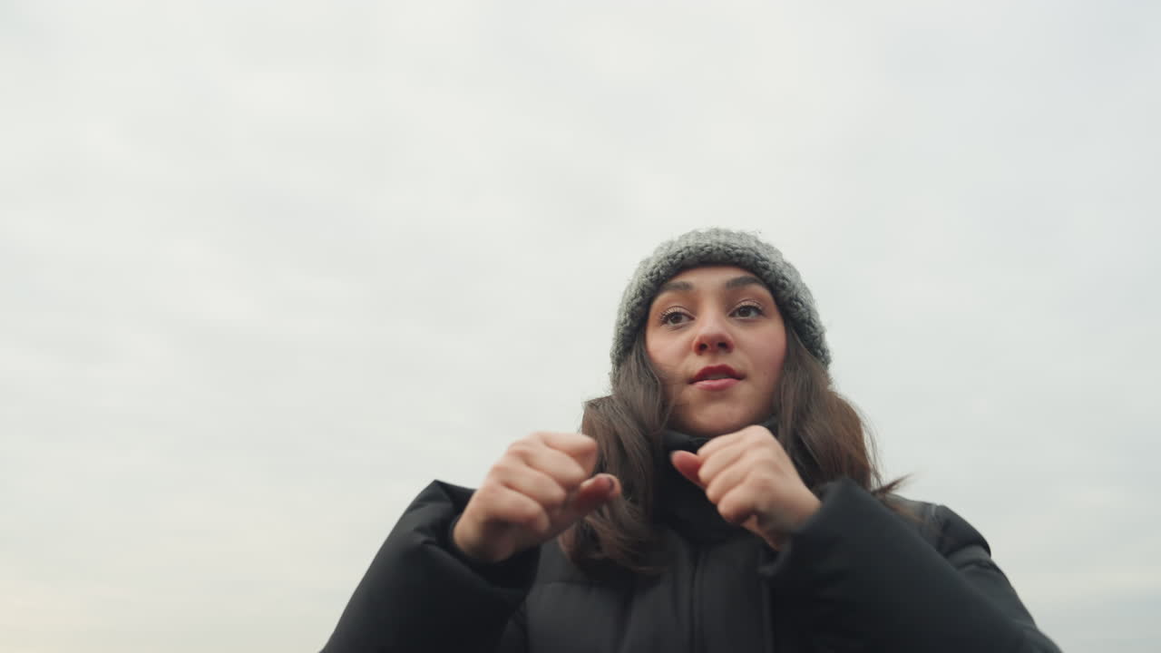 Pratica di dance boxing sul tetto, donna che fa shadowboxing, sfondo dello skyline coperto, berretto di lana e piumino, pugni energici e passi coreografici, allenamento di danzatrice di strada, riscaldamento per l'allenamento fitness.