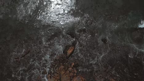 aerial drone view straight down flying over beach with big rocks and boulders as waves come crashing in